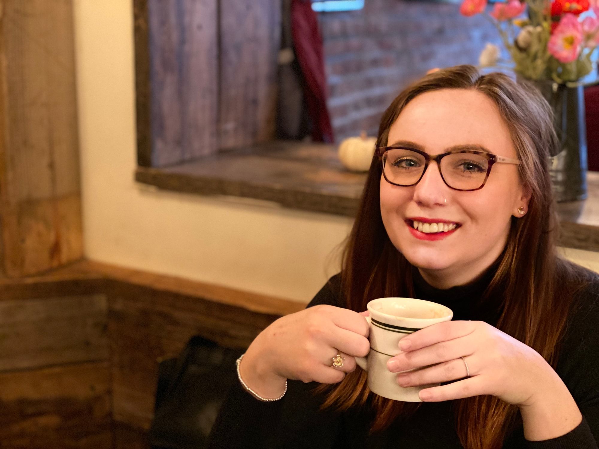 Hannah Isles, smiling, wearing dark-rimmed glasses and a black roll-neck jumper, holding a cup of coffee at a table in a café.
